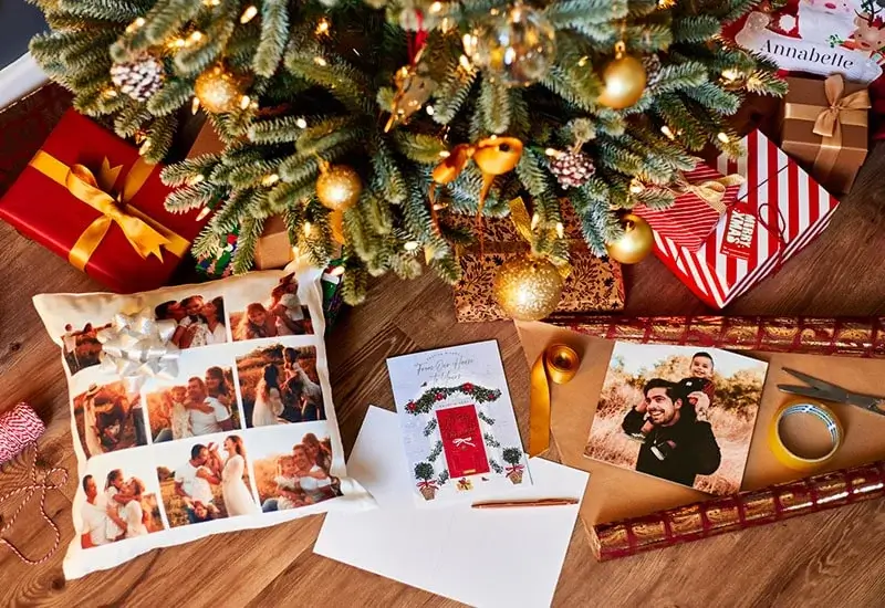 A decorated Christmas tree with gold ornaments and lights surrounded by festive items on a wooden floor, including wrapped gifts, a personalized photo pillow, a holiday card, blank paper with a pencil, a printed photo, wrapping paper, scissors, and tape.